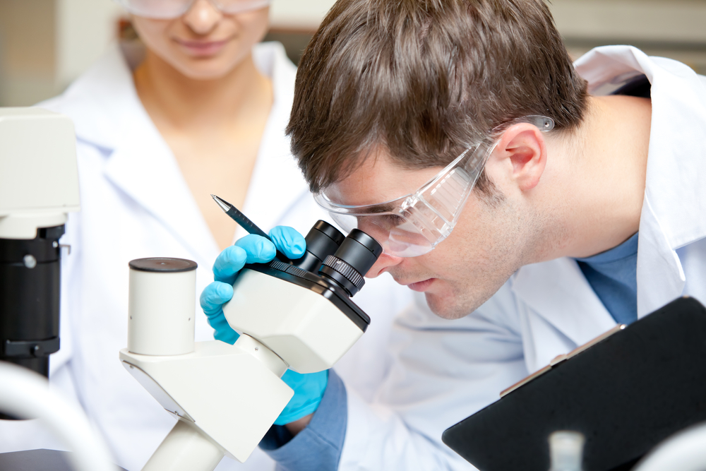 Caucasian male scientist holding pen and clipboard looking through a microscope in his laboratory